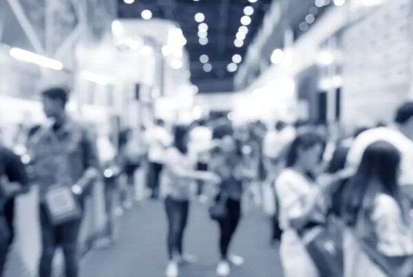 A blurred image of a crowded indoor event with people walking and interacting, showcasing vibrant exhibition booths. The background is filled with bright lights, highlighting the latest in MIM 3D printing technology.