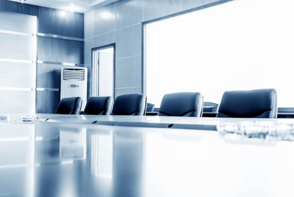 A modern conference room with a long, polished table, black leather chairs, and glass walls, illuminated by bright natural light. An air conditioning unit is visible in the corner.