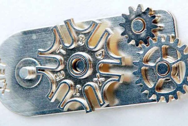 Close-up of a small mechanical assembly with several interlocking gears and a stamped metal plate. One gear is uniquely shaped like a star, and the entire assembly appears metallic and intricate.