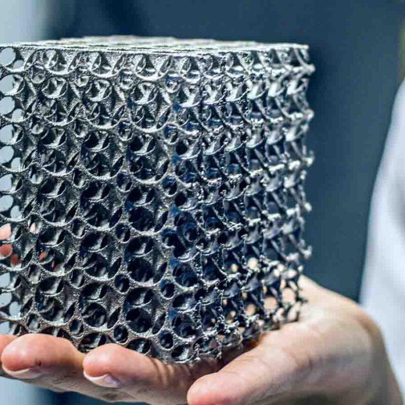 A person in a white lab coat holds a 3D-printed metallic lattice cube with intricate hollow structures, showcasing the tangible results that highlight the pros and cons of 3D metal printing.
