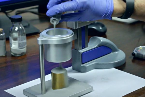 A person wearing blue gloves is pouring liquid from a small bottle into a metal apparatus on a wooden desk with several small bottles in the background.