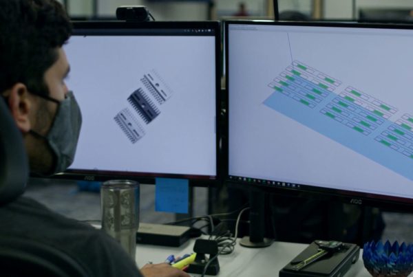 Man wearing a face mask working on two computer monitors displaying technical blueprints in an office setting.