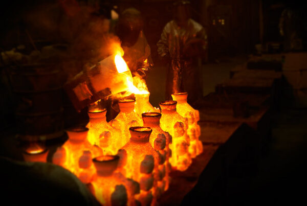 Workers in protective gear pour molten metal into molds, creating a glowing effect in a dimly lit foundry, reminiscent of the advanced techniques that have driven down the cost of 3D printing.