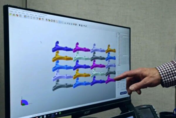 A hand points at a computer screen displaying a 3D modeling software with various colored mechanical parts arranged in a grid.