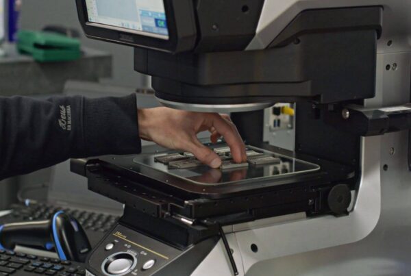 A person operates a machine by placing semiconductor wafers on a tray beneath a screen, in a lab or manufacturing setting.