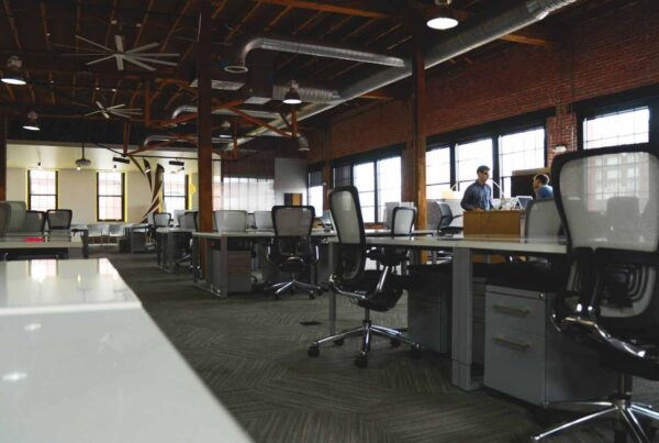 Spacious open office with multiple workstations, ergonomic chairs, industrial design elements, and two people talking in the background near the windows.