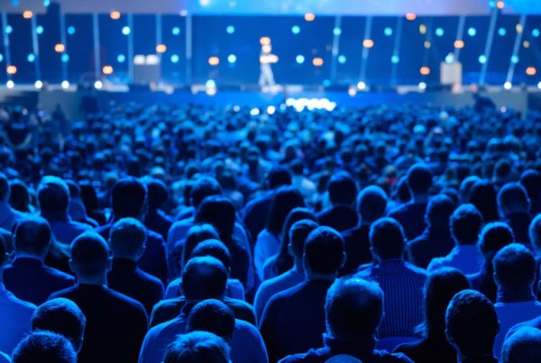 Audience at a conference watching a speaker on stage, illuminated by blue stage lights.