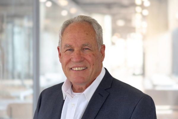 A senior man with a friendly smile, wearing a gray blazer and white shirt, standing in a bright, modern office environment.