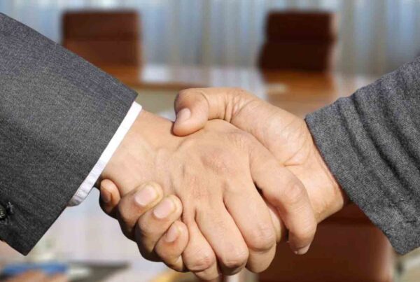 Two individuals shaking hands in a formal setting, with a blurred background of a conference table and chairs.