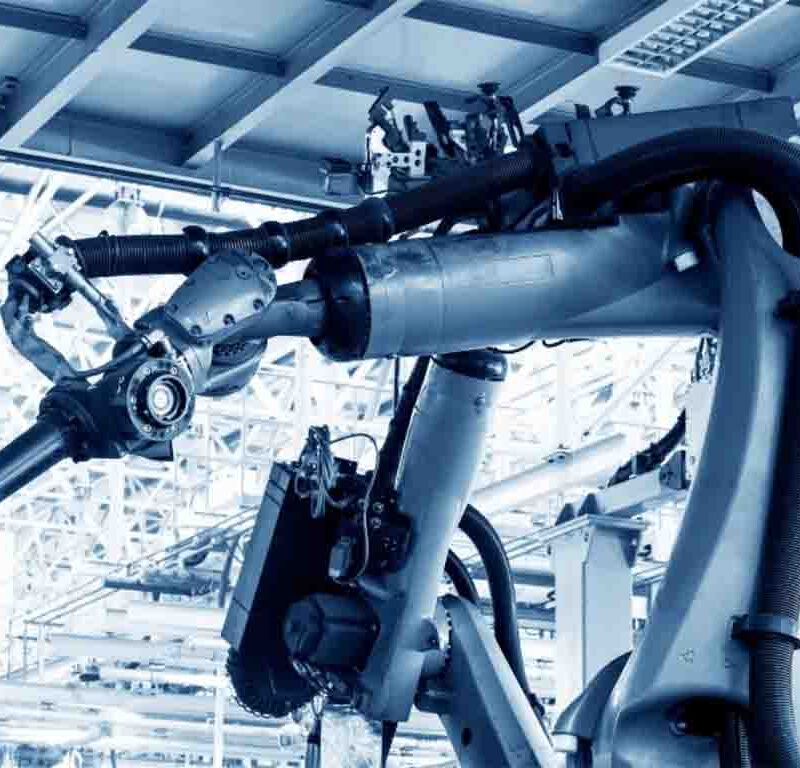 A large industrial robotic arm operates in a modern factory setting with metal beams and machinery visible in the background.