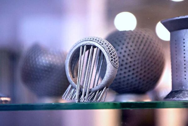 Close-up of intricately designed metallic 3D-printed objects on display, including a sphere with perforations and a ring-like structure with vertical rods. The background is blurred.