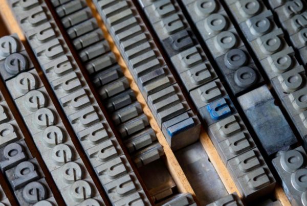 A close-up of metal letterpress type blocks arranged in rows, including letters, numbers, and symbols, typically used in traditional printmaking.