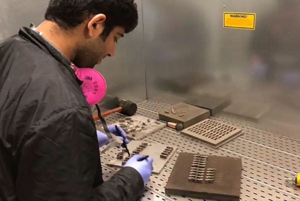 A person wearing a black jacket and blue gloves works on small metallic components inside a lab workstation.