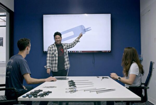 Three people are in a conference room. One person is presenting a CAD drawing of a mechanical part on a wall screen while the other two are seated and listening. Engineering tools are on the table.