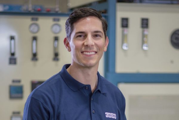 A person wearing a navy-blue collared shirt smiles while standing in front of industrial equipment with gauges and dials.