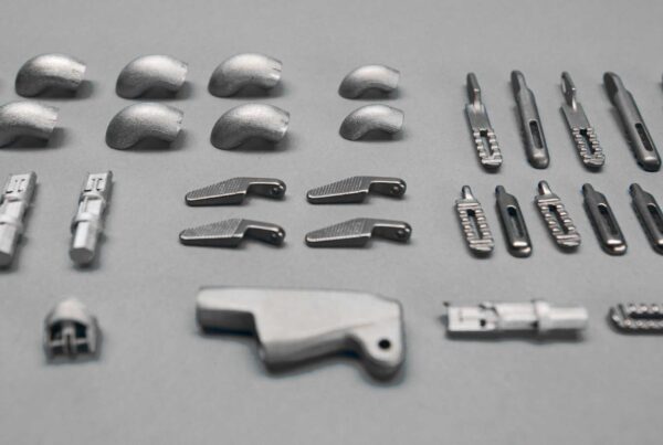 An assortment of metal components is neatly arranged on a gray surface, including curved pieces, connectors, and various small mechanical parts.