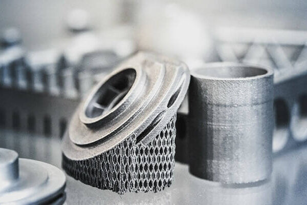 Close-up of several metallic 3D-printed components, including a cylindrical piece and a detailed part with lattice-like structures, on a reflective surface.