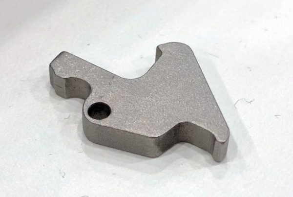 Small metal component with a unique, angular shape and a single hole, resting on a white surface.