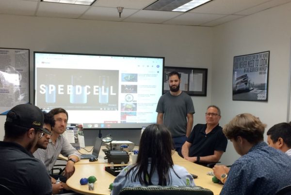 A group of people sit around a conference table during a meeting. A person stands near a screen displaying "SPEEDCELL." The room has posters and a whiteboard on the walls.