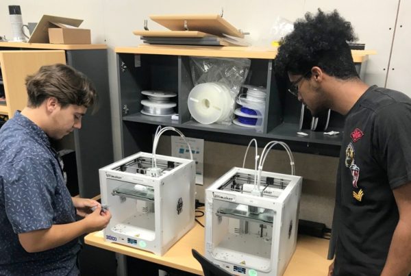 Two individuals stand next to two Ultimaker 3D printers in an office, examining them. Shelving units with various supplies are in the background.