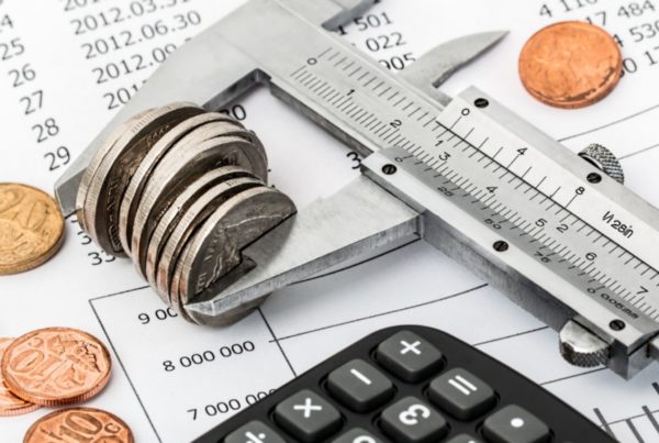 A caliper measures a stack of coins on a financial document, surrounded by additional coins and a calculator.