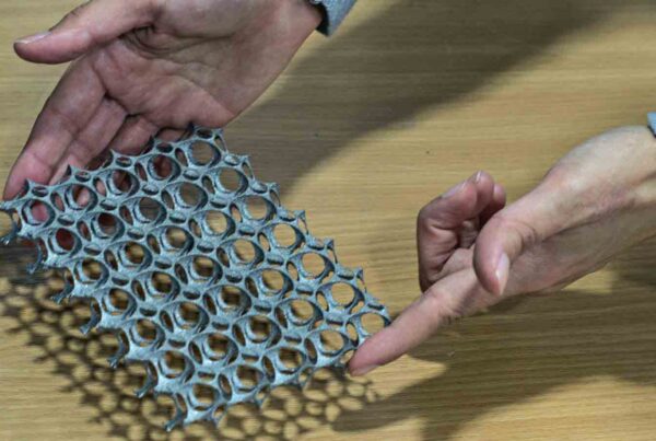 Hands holding and examining a piece of lightweight, flexible, honeycomb-patterned material on a wooden surface.