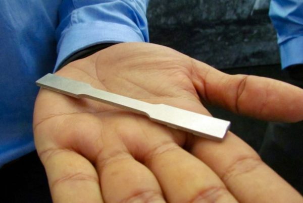 A person holding a metallic key blank produced by a metal 3D printer in their open palm, showcasing it against a blue shirt background.