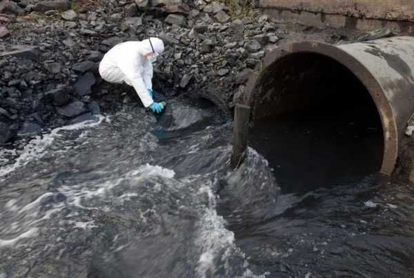 A person in protective clothing collects water samples from a stream flowing out of a large metal drainage pipe.