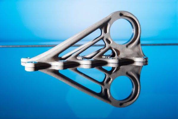 A silver metallic bracket created using 3D metal printing, featuring a complex geometric design, displayed on a reflective blue surface.