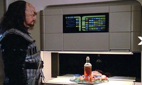 A man in futuristic armor looks at a computer display above a table with food and drinks, showcasing designs from a 3D metal printer.