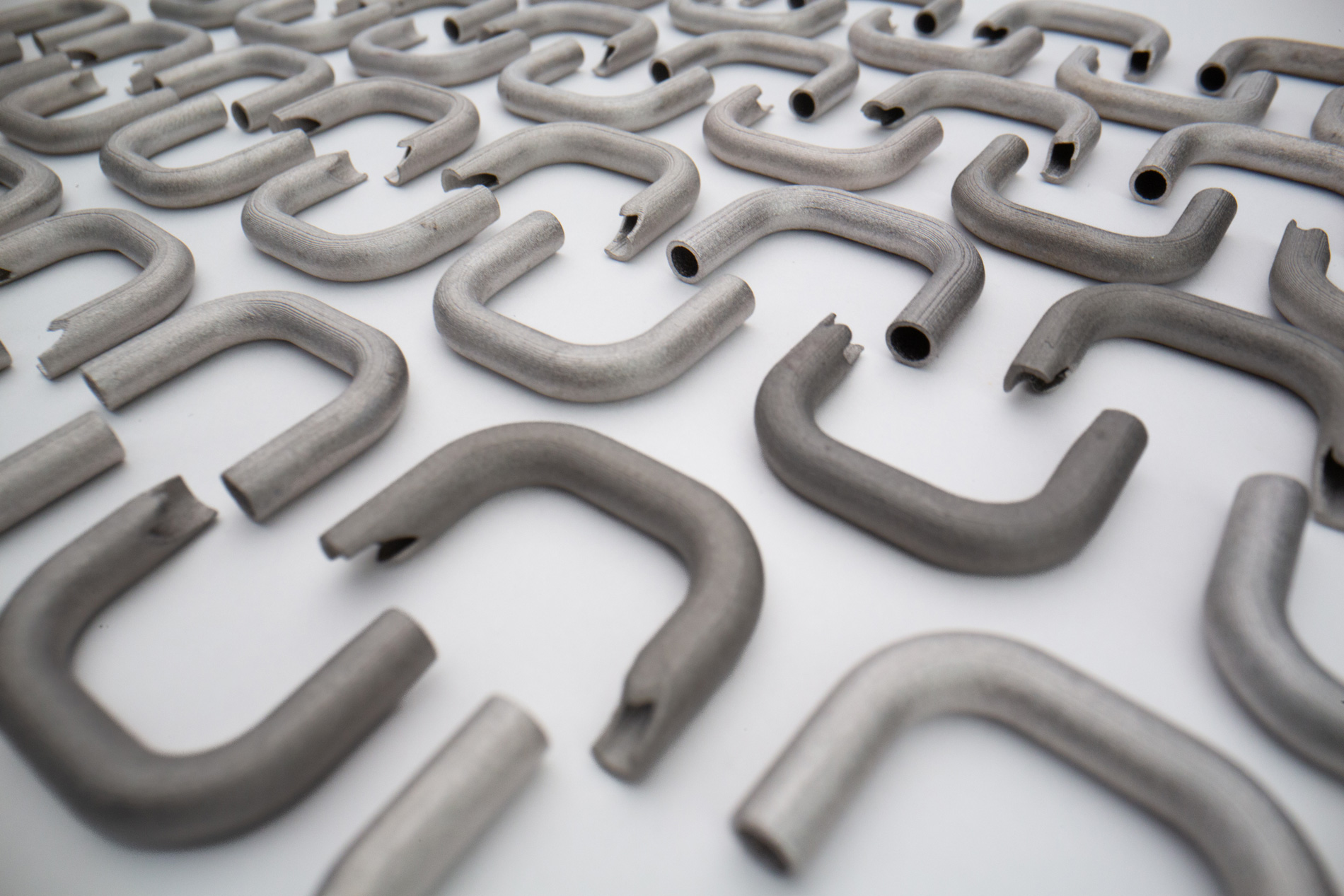 A white surface covered with numerous uniformly arranged, 3D metal printed U-shaped pipe sections with open ends.