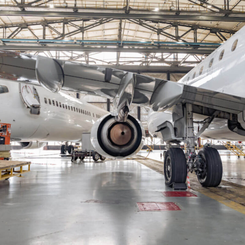 Two white airplanes undergoing maintenance inside a hangar. The view focuses on the landing gear and engines of the aircraft. The area, equipped with scaffolding and various tools, is a hub of aerospace manufacturing precision.