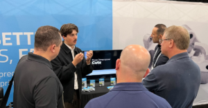 A man is presenting to four other men in front of a display booth at an event. The display includes a TV screen showcasing "Three Dimensional Cuts" related to aerospace manufacturing, and a table with brochures and promotional materials.