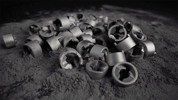 A pile of small, metallic, cylindrical parts, likely produced through 3D printing metal technology, rests on a dusty surface.