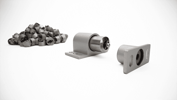 A pile of metal cylindrical objects, possibly products of 3D printing metal technology, lies on the left, while a few individual cylindrical components with mounted brackets are arranged on the right. All are placed neatly on a white surface.