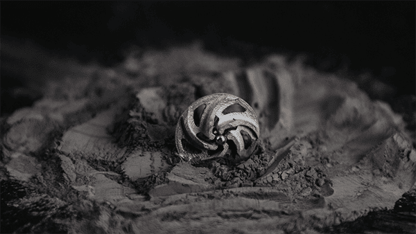 A small, intricately designed metal sphere with geometric cutouts, possibly created through 3D printing, rests in the center of a dusty, uneven surface.