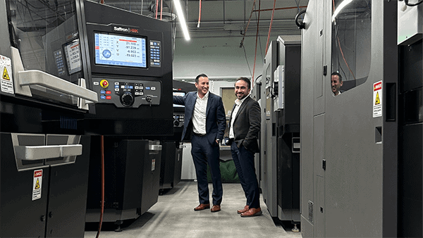 Two men in business attire stand and smile in a modern machine room, surrounded by advanced industrial equipment and control panels, including state-of-the-art 3D printing metal machines.