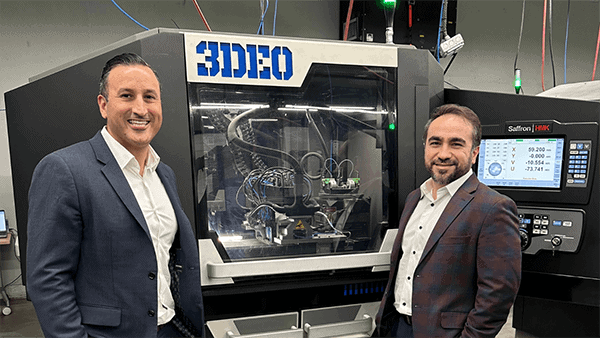 Two men in business attire stand beside advanced 3D printing machinery with the label "3DEO" in a manufacturing facility, specializing in 3D printing metal.
